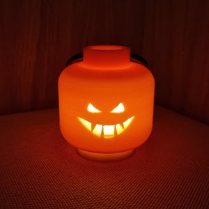 May include: An orange Halloween jack-o'-lantern shaped like a Lego brick with a carved scary face. The lantern is lit from inside, casting a warm glow.