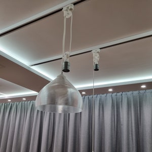 May include: Two pendant lights with silver-colored, bell-shaped shades hang from a white track system. The shades are suspended by white cords. The background features gray curtains and a neutral-toned ceiling with recessed lighting.