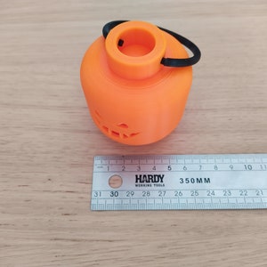May include: An orange 3D printed pumpkin shaped candle holder with a black elastic handle. The candle holder has a jack-o-lantern face carved into the front.