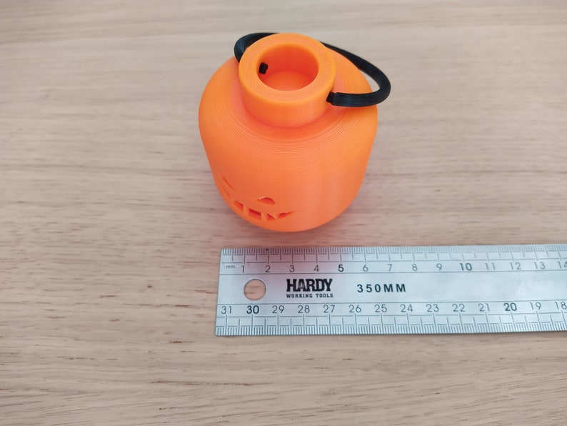 May include: An orange 3D printed pumpkin shaped candle holder with a black elastic handle. The candle holder has a jack-o-lantern face carved into the front.