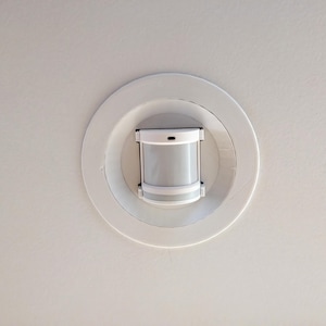 May include: A white motion sensor mounted within a circular white recessed housing. The sensor features a cylindrical, semi-transparent gray lens section with a small black indicator light at the top, centered inside the circular frame.