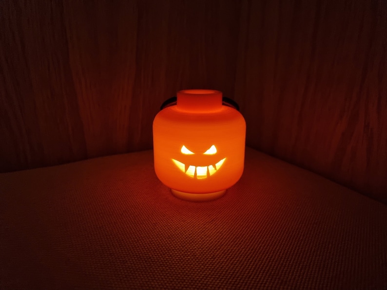 May include: An orange Halloween jack-o'-lantern shaped like a Lego brick with a carved scary face. The lantern is lit from inside, casting a warm glow.