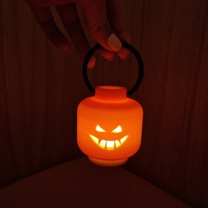May include: A small orange pumpkin-shaped lantern with a black handle. The lantern has a carved jack-o'-lantern face with a lit candle inside.