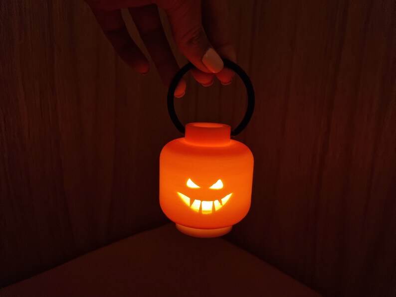 May include: A small orange pumpkin-shaped lantern with a black handle. The lantern has a carved jack-o'-lantern face with a lit candle inside.