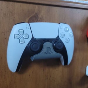 May include: A white PlayStation 5 DualSense controller with black accents is mounted on a wall. The controller has a PlayStation logo on the front.