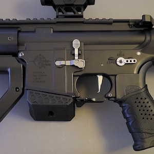 May include: A black and silver rifle with a textured grip and a complex design. The rifle features a scope, a forward grip, and a stock. The metal components are silver, and the rest of the rifle is black.