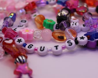 May include: Close-up of colorful beaded bracelets with various shapes and colors. The bracelets include beads with the word "GUTS" spelled out. Other beads are shaped like stars, flowers, and hearts. The color palette includes pink, purple, orange, and white.