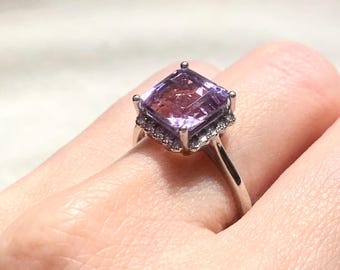 Puede incluir: Anillo de plata con una piedra de amatista tallada en forma cuadrada. La gema púrpura tiene un degradado de color, más oscuro en el centro. Pequeñas piedras claras rodean la amatista, añadiendo brillo a la joya.