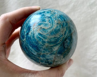 May include: A polished, spherical gemstone in shades of teal and turquoise, with a swirling pattern. The stone is held in a hand, showcasing its smooth, reflective surface. The gemstone's color and texture create a captivating visual effect.