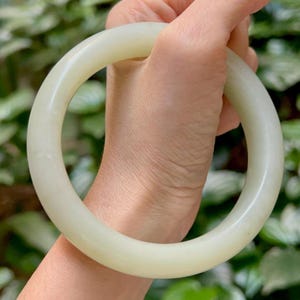 Pode incluir: Uma pulseira circular lisa, feita a partir de uma peça sólida de pedra de jade de cor branco-sujo, pálida e translúcida. A pulseira apresenta uma superfície polida e arredondada com uma espessura consistente ao longo da circunferência.