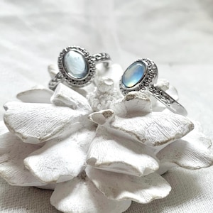 May include: Two silver rings with oval, iridescent gemstones. The rings have ornate, textured bands and are set on a white, textured floral sculpture. The gemstones display a range of colors, including blue and white.