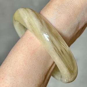 May include: A light green and cream-colored bangle bracelet. The smooth, polished surface of the bracelet displays a marbled pattern. The bangle is a circular shape and appears to be made of a solid material.
