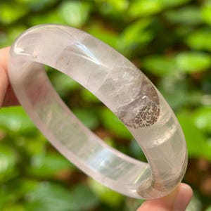 以下が含まれることがあります： ユニークなインクルージョンを持つ透明な水晶バングル。ブレスレットは明るく半透明の色で、茶色の模様が入っています。ブレスレットの滑らかで丸みを帯びたデザインは、ぼやけた緑色の背景に対して見えます。