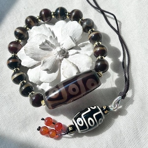 May include: A beaded bracelet and pendant featuring dzi beads. The bracelet has dark brown and tan beads with gold-coloured spacers. The pendant has a cylindrical dzi bead with a geometric pattern, and a black and white bead with a geometric design.