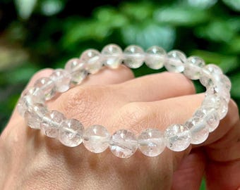 May include: A clear quartz crystal bracelet. The bracelet is made of round, polished beads strung together. The beads are translucent with slight internal inclusions. The bracelet is worn on a wrist, with a blurred green background.