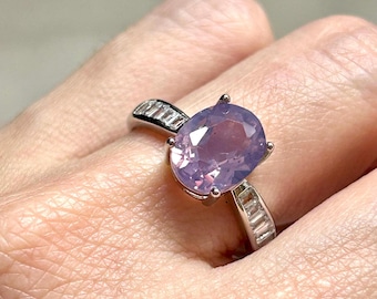 Puede incluir: Un anillo de plata con una gran piedra preciosa ovalada de color púrpura y una banda de piedras preciosas más pequeñas y transparentes.