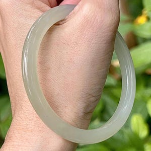 Puede incluir: Un brazalete de jade verde pálido. La superficie lisa y pulida refleja la luz, mostrando las variaciones naturales de la piedra. El diseño circular es un estilo de joyería clásico, adecuado para el uso diario o ocasiones especiales.