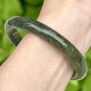 Puede incluir: Un brazalete de jade verde. La superficie pulida refleja la luz, resaltando las variaciones naturales de color. El brazalete tiene un diseño circular y sólido. El jade es de color verde oscuro, con motas más oscuras.