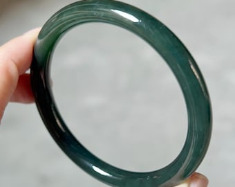 May include: A dark green jade bangle bracelet. The smooth, polished surface reflects light, showcasing the natural variations in the stone. The circular shape is classic and elegant, suitable for everyday wear or special occasions.