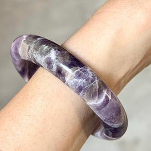 May include: A purple and white amethyst bangle bracelet. The bracelet is made of polished stone and has a swirling pattern.
