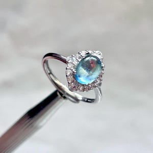 Könnte beinhalten: Ein silberner Ring mit einem ovalen, hellblauen Edelstein, umgeben von kleinen, klaren Steinen. Der schmale, polierte Ring des Rings ergänzt die reflektierende Oberfläche des Edelsteins und erzeugt einen funkelnden Effekt. Der Ring ist vor einem weichen, verschwommenen Hintergrund platziert.