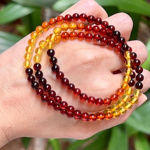 Op de afbeelding: Drie kralenarmbanden in tinten amber, oranje en dieprood worden getoond. De armbanden zijn gemaakt van kleine, ronde kralen en worden in een hand gehouden. De kralen hebben een glanzende afwerking.