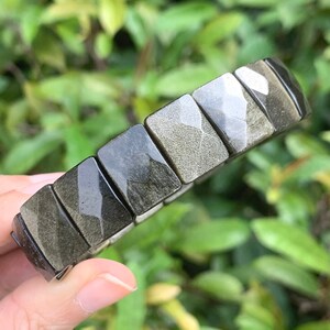 Könnte beinhalten: Ein Armband aus schwarzem und goldenem Obsidian. Das Armband besteht aus rechteckigen, facettierten Perlen. Die Perlen haben einen reflektierenden, metallischen Glanz. Das Armband wird vor einem Hintergrund aus grünem Laub gehalten.