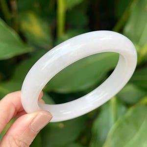 May include: A white jade bangle bracelet. The bracelet is a solid, circular band with a polished finish. The background is a blurred view of green foliage.