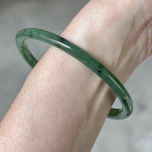 Può includere: Un braccialetto a cerchio in giada di un verde intenso, con sottili inclusioni verde scuro e nere. La superficie liscia e lucida riflette la luce, evidenziando la forma circolare del braccialetto. Un elegante gioiello classico.