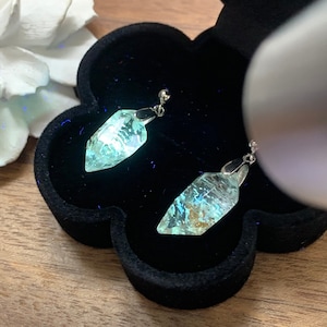 Pode incluir: Um par de brincos de prata com um pingente em forma de cristal azul claro iridescente. Os brincos estão em uma caixa de joias de veludo preto.
