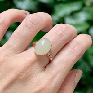 Op de afbeelding: Een roségouden ring met een ovale, lichtgroene edelsteen. De ring heeft delicate details aan de zijkanten van de band, en de edelsteen is gezet in een klassieke prong setting. De ring wordt aan een vinger getoond.