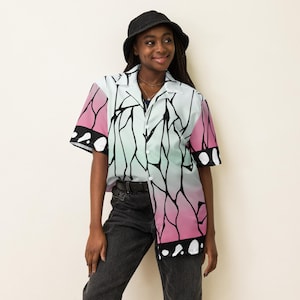 May include: Short-sleeved button-down shirt with a butterfly wing design in pink, white, and light green. The shirt has a black outline pattern and black accents on the sleeves. The model is wearing a black bucket hat and dark jeans.