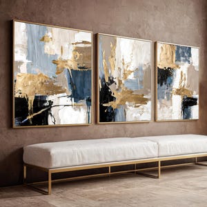 Puede incluir: Tres cuadros abstractos enmarcados con pinceladas doradas, azules, negras y blancas. Cuelgan sobre un banco blanco con marco de metal dorado. Una lámpara de pie con pantalla blanca está cerca.