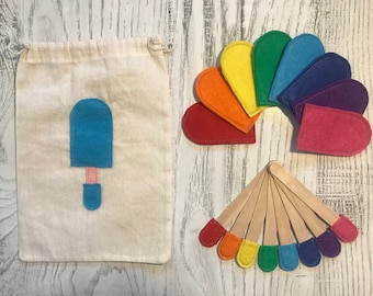 May include: A set of felt rainbow popsicle shapes, a felt blue popsicle shape, and a white drawstring bag. The popsicle shapes are arranged in a semi-circle, with the blue popsicle shape in the center of the bag.