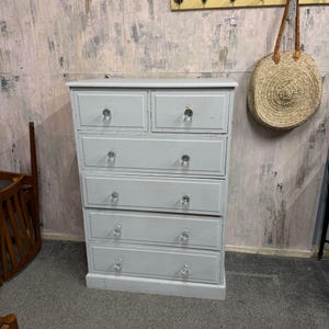 May include: A pale grey wooden chest of drawers with six drawers and crystal knobs. The chest has a rectangular shape and is painted in a solid, neutral colour. A woven straw bag hangs on the wall in the background.