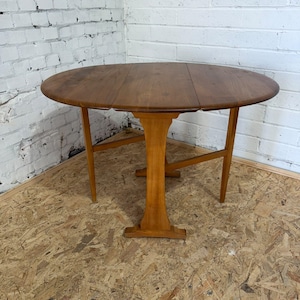 May include: A medium-brown wooden drop-leaf table with an oval top. The table features a central pedestal base with a vertical support column and two hinged side leaves that can be lowered. The wood grain is visible across the surface.