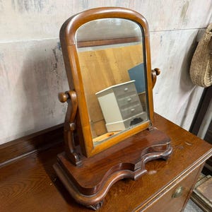 May include: A wooden vanity mirror with a rectangular, arched top. The mirror is framed in a warm, medium-toned wood and is supported by adjustable side arms. The base is a matching wood with decorative curves and carvings. The mirror reflects a room with a chest of drawers.