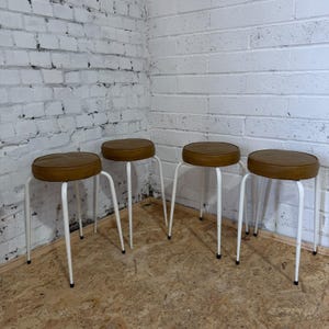 May include: A set of four stools, each featuring a round, mustard-brown padded seat and four slender, white-painted metal legs. The stools are arranged in a row, showcasing a consistent design with simple, clean lines and a functional structure.