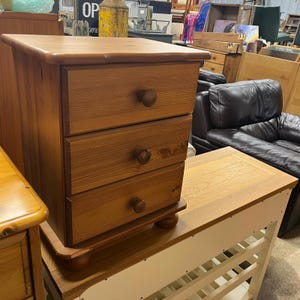 May include: A three-drawer wooden bedside table with a warm, natural finish. The drawers have round wooden knobs. The bedside table is sitting on top of a white and wood table. A classic piece of bedroom furniture.