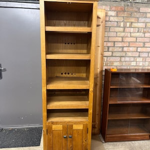 May include: A tall, wooden bookcase in a light oak finish. It features multiple shelves and a cabinet at the base with two doors. The bookcase is empty, offering space for books or decorative items. The height is approximately 180 cm.