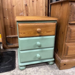 May include: A three-drawer wooden bedside table. The top drawer is natural wood, and the two lower drawers are painted a light green. Each drawer has a wooden knob, and the table rests on rounded feet.