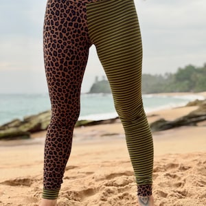 Op de afbeelding: Hoge taille legging met een gesplitst ontwerp: de ene kant heeft een bruine luipaardprint, de andere kant heeft olijfgroene en gele strepen. Het model staat op een zandstrand. De legging is van rekbaar materiaal.