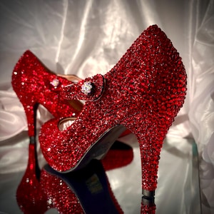 May include: A pair of red high heels covered in sparkling rhinestones. The shoes have a Mary Jane strap with a decorative jewel and a stiletto heel. The shoes are reflected in a mirrored surface.
