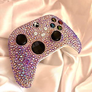 Może przedstawiać: Kontroler Xbox pokryty iryzującymi kryształkami. Kontroler ma czarne joysticki i przyciski, z kolorowymi przyciskami dla X, Y, A i B. Projekt to mieszanka fioletu, różu i złota.
