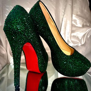 May include: A pair of high-heeled pumps completely covered in small, dark green crystals. The shoes feature a high stiletto heel, a rounded toe, and a distinct bright red sole. The interior lining of the shoes is a light beige color.