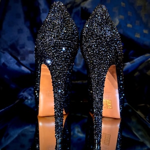 May include: A pair of black high-heeled shoes covered in sparkling rhinestones. The shoes have a pointed toe and a stiletto heel.