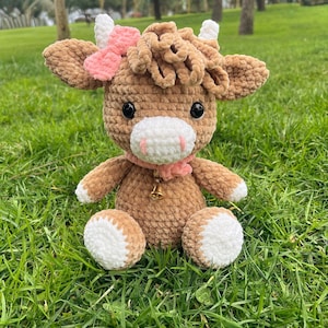 May include: A handmade crochet cow plush toy. The cow is brown with white accents on its hooves and horns. It has a pink bow and scarf, and a small gold bell. The plush is sitting on green grass.