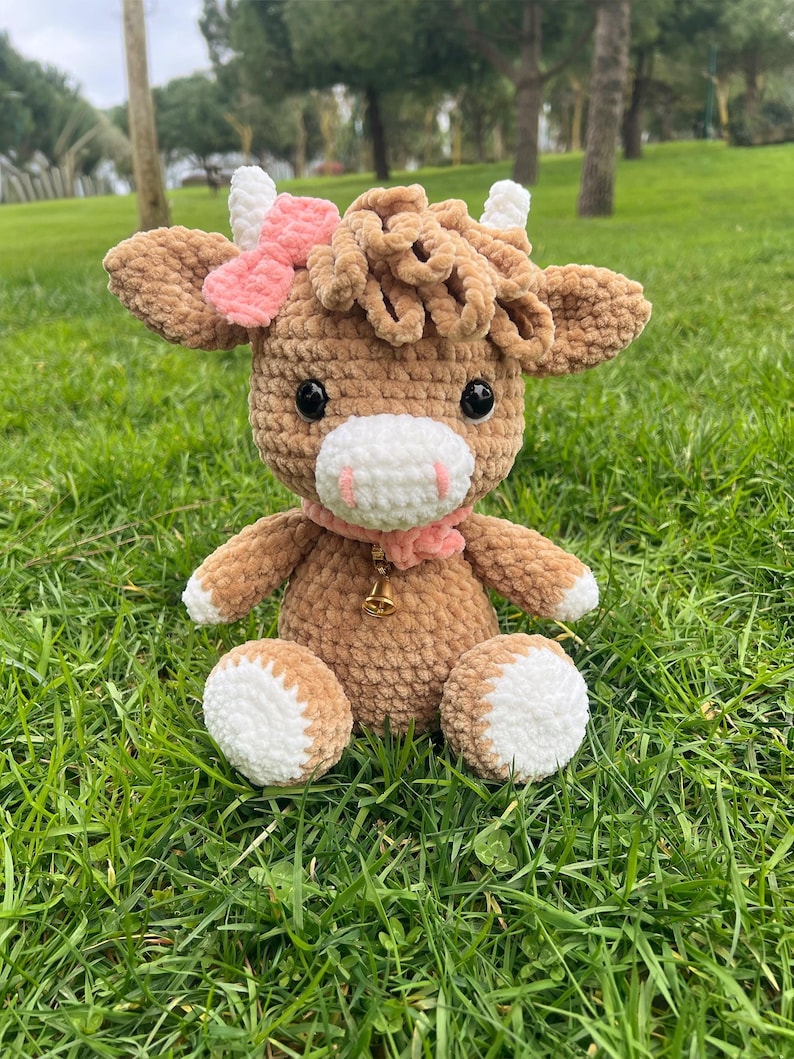 May include: A handmade crochet cow plush toy. The cow is brown with white accents on its hooves and horns. It has a pink bow and scarf, and a small gold bell. The plush is sitting on green grass.