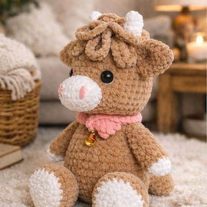 May include: A handmade brown crochet cow plushie with white accents on the face, feet, and horns. It has a pink scarf and a gold bell. The plushie is sitting on a white rug.