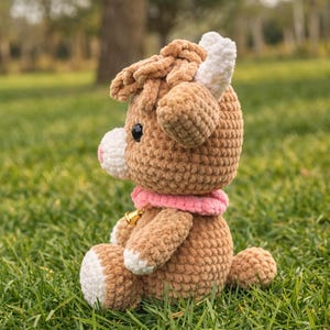 May include: A handmade crochet cow plush toy in shades of brown, white, and pink, sitting on green grass. The plush has a pink collar with a small gold bell. The cow's horns and hooves are white.